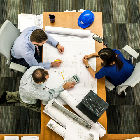 Overhead view of three colleagues at a table, reviewing drawings and costing them out on a calculator